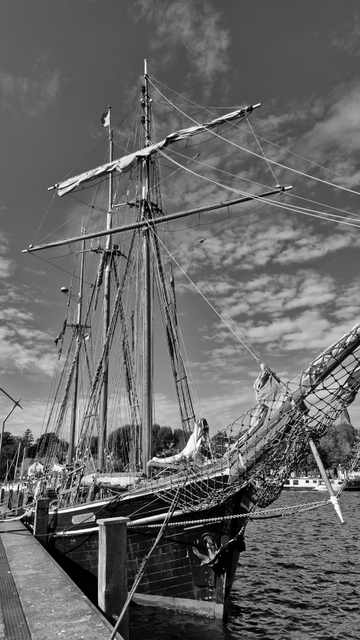 Es handelt sich um eine Schwarzweißfotografie eines Segelschiffs mit drei Masten, das im Hafen am Kai festgemacht ist. Die Aufnahme erfolgte von schräg vorne, sodass ein Teil der rechten Seite des Schiffes zu sehen ist. Auf der Aufnahme ist links der Kai und rechts das Wasser zu sehen. Im Hintergrund ist am gegenüberliegenden Ufer urbane Bebauung zu erkennen. Der Himmel ist leicht bewölkt. 