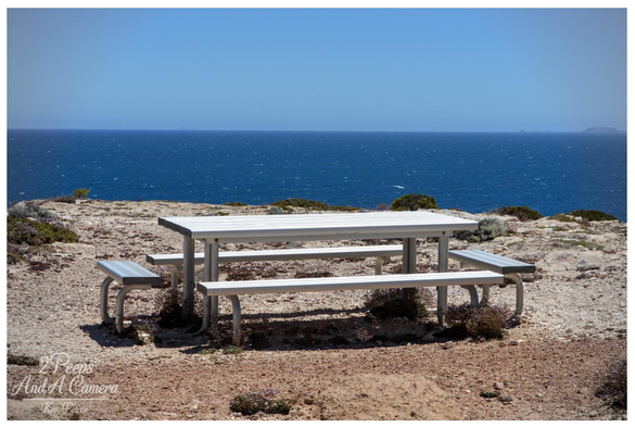 A white metal picnic table and two benches sit on a reddish brown, rugged cliff overlooking a deep blue ocean that meets a bright clear sky.