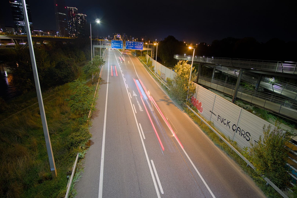 Das Bild zeigt eine nächtliche Autobahn in der Stadt: Die Langzeitbelichtung verwandelt die Lichter der Autos in rötliche Lichtspuren. Rechts neben der Fahrbahn verläuft ein Fuß- und Radweg, an dessen Wand ein Graffiti mit der Aufschrift "FUCK CARS" zu sehen ist. Im Hintergrund ragen beleuchtete Hochhäuser auf, was der Szene eine urbane Atmosphäre verleiht.