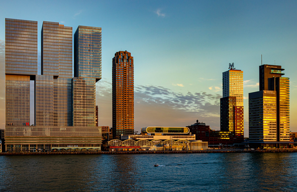 A photo across the river to the other bank, where buildings are illuminated golden by the setting sun. The sky is clear and blue (blue hour). At the bottom, a horizontal stripe of water, dark blue due to the reflection of the sky. On the opposite bank, along the river to the left, a very tall and very wide building, which appears to be made of large, stacked rectangular blocks. Next to it, a slightly lower, narrow tower of dark red stone. In the middle, low-rise buildings, followed by another group of tall towers, golden in the sun.