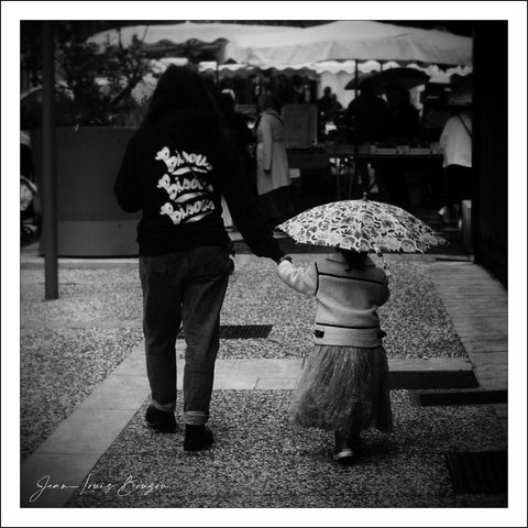 This black-and-white image evokes a tender and timeless moment between two figures walking away from the camera down a pathway. The contrast between light and shadow brings out the textures of the pebbled ground and the soft flow of the little girl's skirt. The taller figure, possibly an older sibling or parent, wears dark pants and a hoodie with a bold, expressive script on the back, which stands out against the monochromatic palette.
The child, by contrast, carries a patterned umbrella that offers a delicate detail, almost like a protective shield against rain or sun, adding an element of softness and whimsy. Holding hands emphasizes their close bond and the theme of guidance, safety, and companionship. The blurred background suggests a lively, possibly outdoor market or social setting, but the focus remains intimate and personal.
The use of black and white can symbolize nostalgia or a captured memory, turning an everyday scene into something more poetic—an exploration of protection, childhood innocence, and connection through simple, shared moments. The visual interplay between light and dark enhances the emotional weight without the distraction of color, allowing the viewer’s imagination to fill in the atmosphere and the mood.

