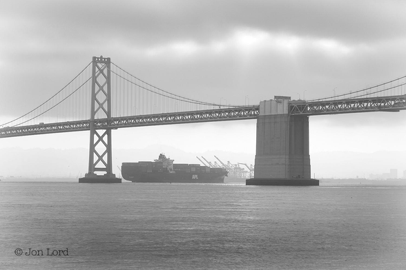This is a black and white photo in landscape format of a large container ship passing under the San Francisco-Oakland Bay Bridge and approaching the port of Oakland, California (2015).

Rising up from the base to one-third up in the image is a body of dark and smooth water. Stretching from the left margin through the centre to the right margin is a long and large suspension bridge. Between the left margin and the centre is one of the bridge's tall towers with the two pairs of suspension cables arching downwards and along to the ends of the deck with the multiple vertical suspender cables descending directly from the from the arching suspension cables to the deck. To the right of centre is an large concrete anchorage that supports the centre of the western section of the bridge, from there the deck continues to the right edge of the image. Framed between the tower and the concrete anchorage is a large container ship, viewed in profile, and underway from left to right. Ahead of the ship are numerous raised gantry cranes in the port awaiting the ship's arrival. The view of the distant shore is slightly obscured by a thin morning mist or haze. Above is an overcast sky, with just a few small breaks allowing the sun to shine through.