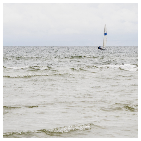 En fyrkantig bild på en liten katamaran på havet med två män. Seglet är vitt med ett blått streck. De är på väg mot land. Himmelen är täckt med lätta moln.