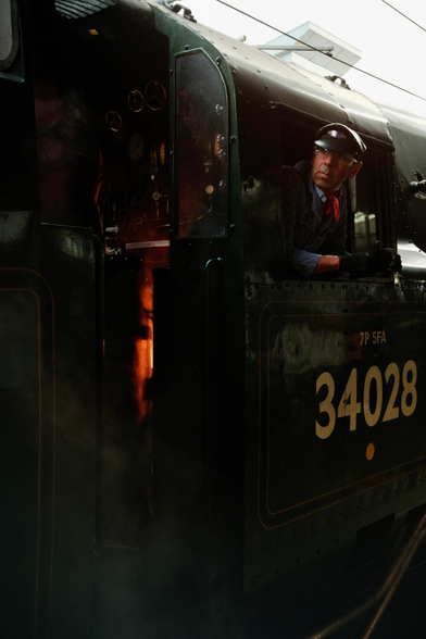 A man wearing a cap leans out of the window of a steam train. Behind him you can see the glow from the fire powering the train.