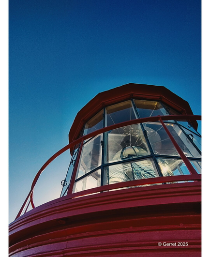 A close-up of a red lighthouse top with glass windows, set against a clear blue sky. The structure's bold lines create a sense of strength and guidance.