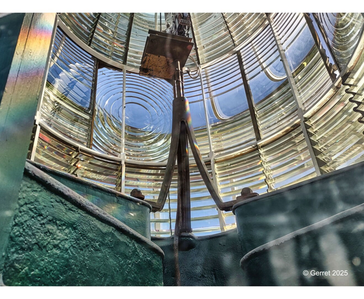 View from inside a lighthouse lens, showing large concentric glass circles and metal framing. The light refracts, creating rainbow effects.