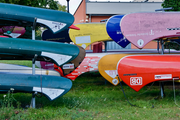 Circa 10 Kanus stehen aufgebockt an Land. Sie hängen kopfüber auf Metallgestellen. Fast alle Kanus haben unterschiedliche Farben und Sponsorenaufdrucke. Auf dem Bild ist nur jeweils eine Spitze der Kanus zu sehen. 