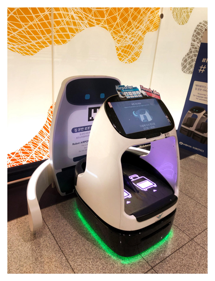 Photo of a robotic luggage carrier docked at its station at an airport, in front of a wall with colorful abstract designs. The carrier has touchscreens and luggage icons and an illuminated base that emits a green glow. The station displays a sign with instructions in Korean and English.