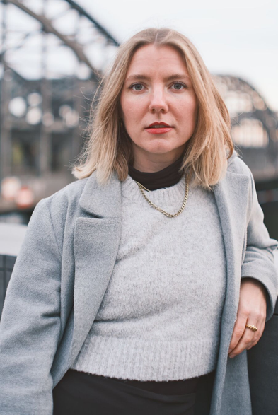 DE: Porträt einer blonden Frau mit grünen Augen. Sie trägt ein grauen Mantel, einen hellgrauen Pullunder und eine goldene Halskette. Ihr Blick geht direkt zur Kamera. Im Hintergrund sind Metallträger einer Brücke zu erkennen.

EN: Portrait of a blonde woman with green eyes. She is wearing a grey coat, a light grey slipover and a gold necklace. She is looking directly at the camera. Metal girders of a bridge can be seen in the background.