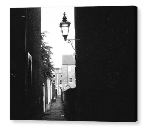 Gloomy black and white photograph looking along a dark alleyway.  The right side is taken up with a dark brick wall on which is affixed a Victorian coaching lamp.  The image is shown printed upon a block canvas.
