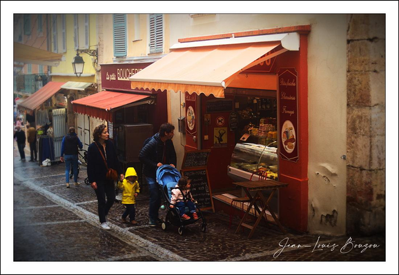 This image captures a charming street scene in what appears to be a quaint European town, possibly in France given the design and language hints from the storefront. The focus centers on a warmly lit storefront with an inviting orange awning that extends over the sidewalk, sheltering a display case filled with delicious treats, likely pastries or ice cream. The shop’s façade is painted in harmonious earthy tones, providing a cozy and welcoming ambience.
Along the cobblestone street, a family strolls peacefully. An adult pushes a stroller holding a content child, while a toddler in a bright yellow raincoat stands nearby, their vivid color contrasting strikingly with the more muted tones of the surroundings. This splash of yellow draws the eye and adds a touch of warmth and cheerfulness to the scene.
The street is flanked by similar buildings — covered shop windows and awnings seem to line both sides, creating a feeling of depth and continuity. Further along, more pedestrians are casually walking, dressed in coats and scarves, suggesting cool or damp weather.
The cobblestones glisten underfoot, likely from recent rain, which adds a reflective quality to the ground that enhances the intimate, cozy feel of the setting. The overall mood is one of leisurely enjoyment and simple pleasures, invoking the timeless charm of browsing local shops on a cool day.
