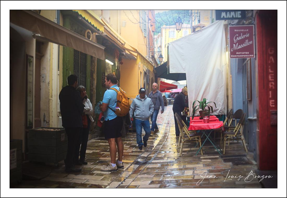 The image captures a narrow, cobblestone street bathed in soft, natural light filtering through the overhead canopy of a quaint European alley. The street is lined with mustard-yellow buildings that evoke a warm, welcoming atmosphere, their walls gently textured and showing signs of age, adding character to the scene. The pathway glistens as if it has recently rained, with reflections softly shimmering on the stones.
On the left, a small group of people are engaged in quiet conversation, their casual attire suggesting a relaxed, perhaps tourist-friendly setting. The figure in the center stands out in a blue T-shirt and black shorts with a bright orange backpack, adding a pop of color that contrasts with the earth tones around him.
To the right, a small café setup is visible—tables covered with red cloths, a single plant adding a touch of greenery, and folding chairs arranged for patrons. The café's inviting but understated appearance blends perfectly with the street's intimate vibe. Above, signs in French hint at the cultural setting, suggesting this could be a charming street in a French city or town.
The overall atmosphere is one of quiet sociability and timeless charm, inviting a slow, mindful exploration. The interplay of light, texture, and human presence evokes a sense of everyday life steeped in history and culture—typical of many European old towns where modern life and heritage coexist seamlessly.
