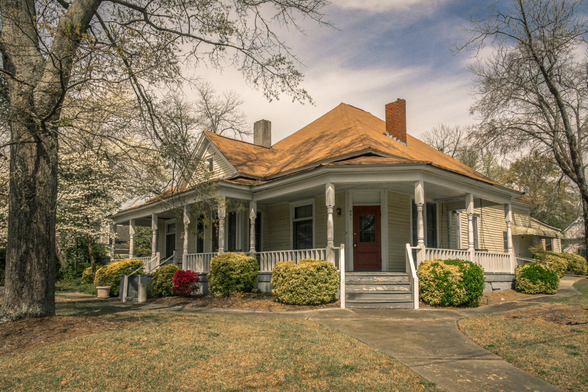 Single-story craftsman-style bungalow with pale green horizontal siding and a wraparound porch supported by white columns and railings. The house features a prominent hipped roof with warm orange-brown shingles and two chimneys - one brick and one cement. A dark wooden front door is centered on the facade, flanked by windows with dark frames. Wide wooden steps lead up to the porch from a concrete walkway. The front yard is landscaped with rounded yellow-green shrubs positioned symmetrically along the porch foundation, and mature bare deciduous trees frame the property on both sides. The sky shows dramatic clouds, creating atmospheric lighting on the home's exterior.