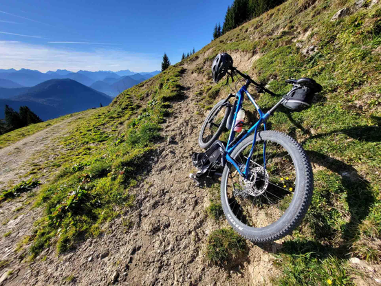Perched on the edge of a rugged mountain trail, a sturdy blue mountain bike takes a well-deserved rest against the backdrop of an endless alpine panorama. The bike, equipped with wide, grippy tires and a sleek black frame bag, leans casually against the slope, its handlebars turned slightly as if gazing out at the view. The trail itself is a mix of sun-baked dirt and scattered rocks, winding through patches of vibrant green grass and wildflowers that dot the hillside.

Beyond the immediate foreground, the landscape opens into a sweeping vista of rolling mountain ridges, their deep blues and purples fading into the horizon under a sky of brilliant, cloudless azure. The crisp, clean air carries the faint scent of pine and wild herbs, while the distant peaks stand silent and majestic, their slopes covered in dense forests. The atmosphere is one of exhilarating solitude—just the trail, the bike, and the boundless beauty of nature stretching out in every direction. It’s a moment frozen in time, capturing the thrill of adventure and the serene reward of reaching such a breathtaking height.