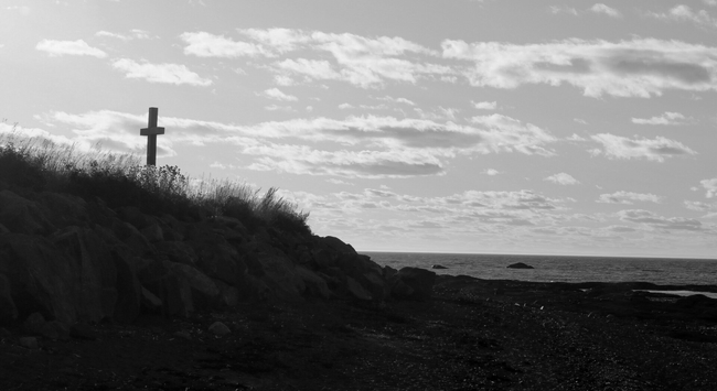 A black and white photograph of a rocky and pebbly beach, with the sea visible to the right stretching to the horizon, under a sunny sky with numerous small clouds. To the left, the rocky side of a higher plot of land is visible, with grass and wildflowers growing along the edge, and a large wooden cross standing against the sky.

Photographie en noir et blanc d'une plage de roches et de galets, avec la mer visible à droite jusqu'à l'horizon, sous un ciel ensoleillé avec des nombreux petits nuages. À gauche, le côté enroché d'un terrain situé plus haut est visible, avec de l'herbe et des fleurs sauvages poussant en bordure, et une grande croix en bois se dressant contre le ciel.