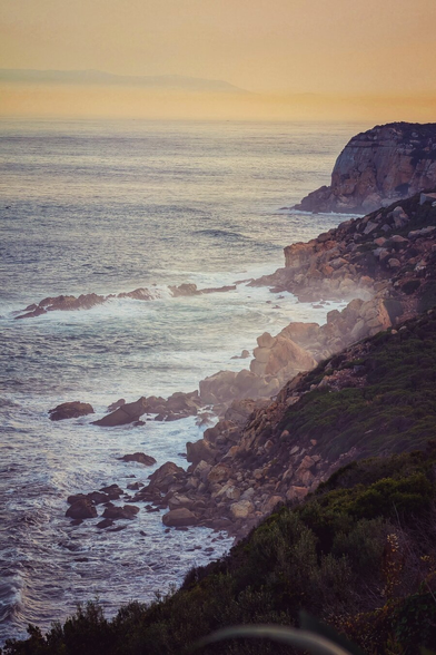 A high-angle photograph of a rugged coastline at either sunrise or sunset. The water is a dark blue-grey, with white waves crashing dramatically against large, dark brown and yellow-hued rocks. The coastline rises to the right, showing a steep, rocky cliff face topped with low, dark green scrub. The sky is a soft blend of pale gold and muted pink, creating a warm, hazy glow over the water and distant horizon. The overall mood is dramatic and serene.