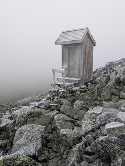 An outhouse standing alone on the loose rocks right at the precipice of the top of a mountain. The rocks and wooden outhouse are completely covered in a thick frost, and there is a thick fog so that the entire picture is an eerie grey.