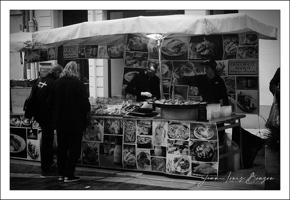 
This black-and-white image captures a small, street-side food stall with a canopy overhead. The stall is covered in numerous photographs of various dishes, creating a visually rich collage that advertises the culinary offerings. The person behind the stall appears to be serving or preparing food, highlighting the active nature of this street food setting.

The two customers in front of the stall add a human element, suggesting interaction and casual dining. Their posture conveys attentiveness, indicating they might be deciding what to order or waiting for their meal.

The monochromatic tone emphasizes textures and contrasts rather than colors, giving the photo a timeless, documentary feel. The array of food images may symbolize cultural diversity or the appeal of street food as accessible, varied cuisine.

Overall, this scene evokes the bustling yet intimate atmosphere of urban food markets, where flavors and human connections intermingle seamlessly.