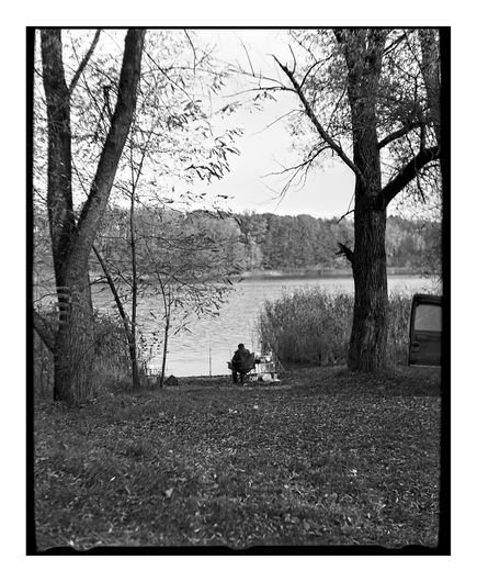 A black and white square film photograph of a person fishing. The person is sitting in a chair on the bank of a lake, framed between two large, leafless trees. The ground is covered in autumn leaves.