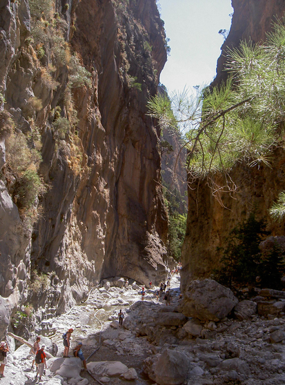 People hiking through a rocky gorge with tall, steep cliffs and sparse vegetation.