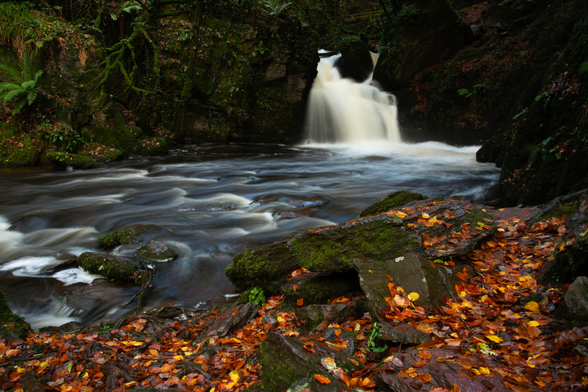 A photo of a waterfall with rocks either side of the fall and more rocks with autumn coloured leaves in the foreground. The stream heads off to the left. The water is blurred by a long exposure.