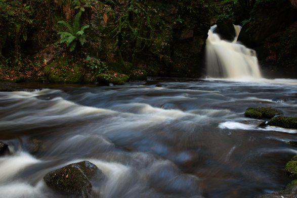 A photo of a waterfall with rocks either side of the fall. The stream heads off to the left and there are more rocks in the stream. The water is blurred by a long exposure.
