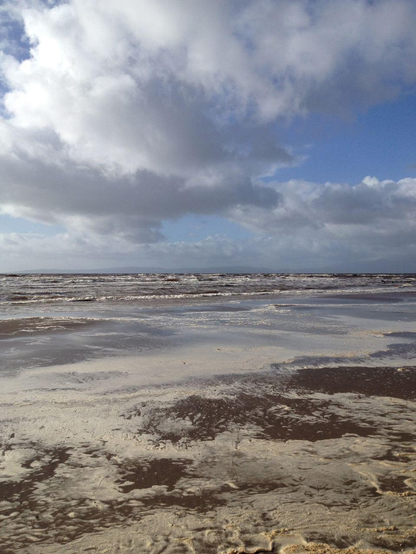 View of the sea with choppy waves and white breakers, and the beach covered in foam. Lively clouds in the sky, leaving quite a bit of blue.