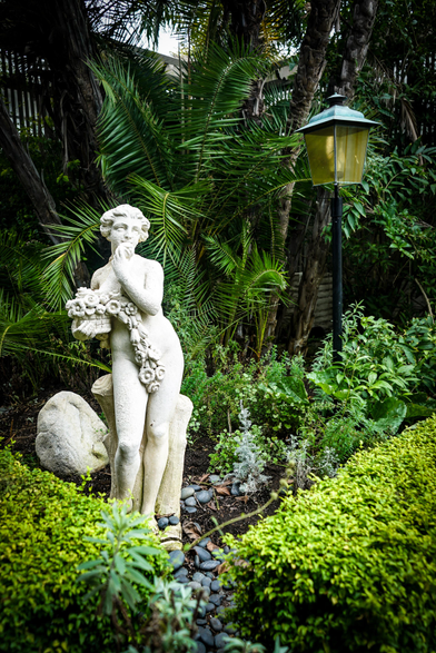 A garden scene features a classical-style statue as a focal point, surrounded by lush greenery. The statue, sculpted in a pale, stone-like material, depicts a figure holding a bunch of what appears to be grapes. It stands amidst dense plantings of various shrubs and low-lying plants with varying shades of green foliage. To the right of the statue is a black lamppost with a yellow-lit lantern. The background consists of more vegetation, including palm fronds, creating a backdrop of rich, dark green hues and a slightly shadowed effect.

Provided by @altbot, generated privately and locally using Gemma3:27b