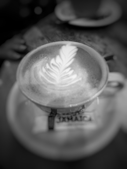 Imagen en blanco y negro de una taza de café con arte latte en forma de hoja o pluma, vista desde arriba. La espuma blanca y clara del café contrasta con el líquido más oscuro de abajo. La taza está sobre un platillo blanco, y debajo de ella se puede ver un paquete pequeño, posiblemente de azúcar, con la palabra 'JAMAICA' parcialmente visible. El fondo está desenfocado, mostrando una mesa de madera oscura y, en la parte superior izquierda, la silueta borrosa de una mano, sugiriendo un ambiente de cafetería.
