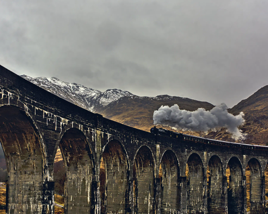 Das aus den Harry-Potter-Filmen bekannte Viadukt spannt sich über das Glen Finnan in Schottland. Soeben fährt der berühmte Jacobite Express dampfend und pfeifend über das Viadukt. Im Hintergrund die Berge Schottlands, zum Teil noch von Schnee bedeckt. Der Himmel ist grau, und es regnet in Strömen.