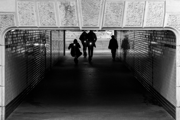 In the black and white photo, three people on scooters are speeding through an underpass. On the right, a pedestrian is walking in the opposite direction. The wall of the underpass is covered with subway tiles, the light glistening on its shiny surface.
