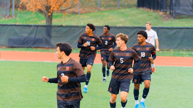 5 soccer players wearing brown uniforms with black stripes are running towards us and and mostly having numbers visible: 3 (i think), 5, 18, 16, 2 with another player with a white jersey in the far distance.  everyone is looking to the right (their left) and there is a red running track and a dark fabric covered fence in the background and past that a grassy field and the bottom of a yellow fall color tree