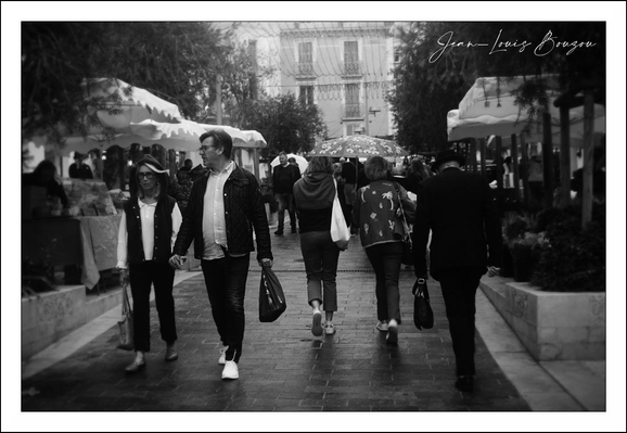 This black-and-white photograph captures a lively street scene filled with pedestrians moving in different directions. The monochrome tone lends a timeless, classic quality to the image, emphasizing contrasts of light and shadow.
In the foreground, two pairs of people walk side by side. On the left, a woman and a man appear engaged, perhaps in conversation, with the woman smiling gently. The man beside her has a casual yet confident stride, carrying a bag in his left hand. They stand out with their lighter clothing, especially the man's open jacket and white sneakers that pop against the cobblestones.
To the right, another pair strolls away from the camera. One wears a star-patterned jacket, which draws attention with its boldness even without color. The other is dressed more formally, possibly in black, suggesting a dynamic contrast not only in style but in mood or activity.
In the middle distance, a third individual—a woman—walks alone with a scarf wrapped around her neck. Her casual yet purposeful walk contrasts with the more social groups around her.
The background reveals an urban setting with trees and traditional architecture, characteristic of European city streets, lined with vendor stalls or market booths under small canopies. This suggests a market or fair atmosphere, inviting community interaction and commerce.
