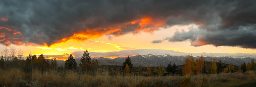 A panoramic view of a vibrant sunset with lively clouds, snow capped mountains in the background, and autumn colored trees and grasses in the foreground.