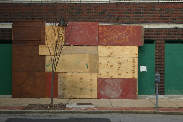 The facade of an old, street-level building. Its doors and windows are boarded-up, covered with plywood painted in red and green. Some sheets remain unpainted. A small, bare tree occupies the left foreground, and a parking meter sites in the right foreground.