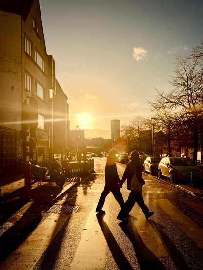Two figures walk hand-in-hand across a zebra crosswalk at golden hour, silhouetted against a bright sun that casts long shadows on the wet street. A building stands on the left, a scooter is parked, and cars line the right side with bare trees. The vibrant sunlight creates strong contrasts and lens flare, illuminating a distant cylindrical building. The scene evokes a warm, atmospheric mood with deep shadows and bright light.