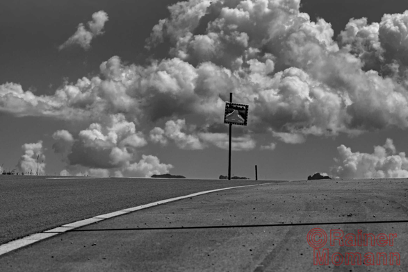 Schwarzweißfotografie im Format 3:2 Landscape.
Eine surreale Asphaltlandschaft mit kraftvollen Kumuluswolken. Im unteren Drittel steigt, die Bildbreite einnehmend, eine asphaltierte Straße an. Eine durchgezogene Fahrbahnmarkierung trennt den Straßenbelag, von links, zur Bildmitte hin. Dieser Schnittpunkt wird von einer Stange, an welchem Ende, ein Spiegel montiert ist, markiert. Darin spiegelt sich eine außerhalb des Bildes, von rechts kommende Straße. Der Himmel über dieser Szene ist mit kräftigen, kontrastreichen Kumuluswolken übersät.