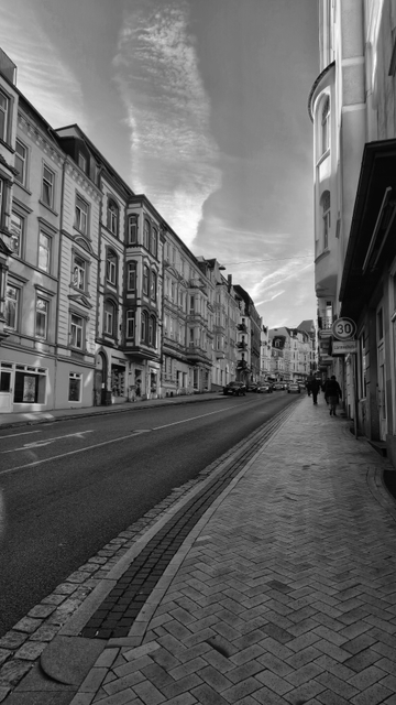 Schwarz-Weiß-Fotografie eines Straßenzugs in Flensburg. Die Aufnahme entstand vom rechten Gehweg aus entlang der Straße mit Schwerpunkt auf die linken Seite. Auf beiden Seiten der Straße befinden sich alte Bürgerhäuser. Davor verlaufen die Gehwege, auf denen vereinzelt Leute zu sehen sind. In der Mitte befindet sich die Fahrbahn, auf der Autos fahren oder am Rand parken. Über den Häusern ist ein leicht bewölkter Himmel zu sehen. 

Black-and-white photograph of a street in Flensburg. The picture was taken from the right-hand pavement along the street, focusing on the left-hand side. There are old town houses on both sides of the street. In front of them are the pavements, where a few people can be seen. In the middle is the road, where cars are driving or parked at the kerbside. Above the houses is a slightly cloudy sky. 