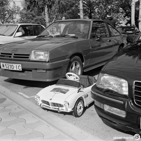 A lineup of cars in black and white, featuring a vintage red sports car and a classic black sedan, with a small toy car parked in front. The scene captures a nostalgic automotive display.