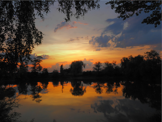Wir sehen einen Sonnenuntergang an einem kleinen See. Das Wasser ist ruhig und so spiegeln sich die Bäume und Sträucher an dessen Rand auf der Wasseroberfläche, wie auch der farbige Himmel. Das Foto wird von Ästen im Vordergrund eingerahmt. Die Sonne selbst ist schon untergegangen und nur noch ihre gelb-orangenen Farben sieht man am Horizont.