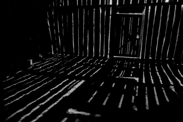 A black and white landscape photo from the inside of an old barn. There are large gaps in the vertical wall planks that casts a striped pattern of sunlight on the ground. 