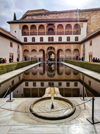 A symmetrical view of the Court of the Myrtles at the Alhambra palace in Granada, Spain. The image captures a long, reflective pool flanked by neatly trimmed hedges, leading to an ornate, historic building with intricate arches and balconies. The building features a series of arches on both the ground and upper levels, showcasing traditional Islamic architecture. At the centre of the pool, there is a small, circular fountain with water gently flowing. The sky above is partly cloudy, and visitors can be seen exploring the courtyard in the background. The overall scene is serene and architecturally stunning, highlighting the harmony between water, architecture, and nature.