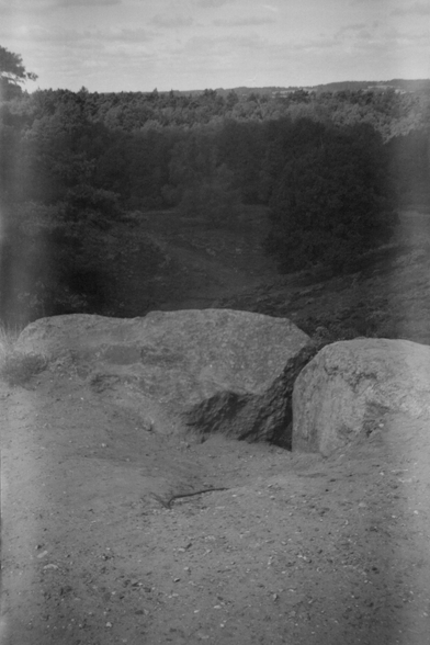 Ein schwarzweißes Landschaftsfoto. Im Vordergrund ist ein Sandweg mit zwei großen Steinen zu sehen. Der Blick geht in eine weite bewaldete Hügellandschaft mit bewölktem Himmel.