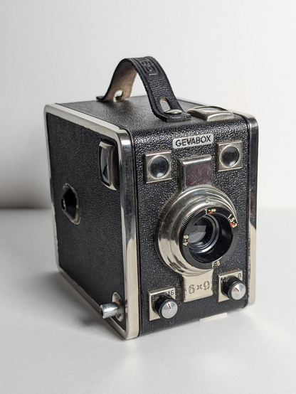 Eine Boxkamera "Gevabox" aus dem Jahr 1954. Vorn ist das Objektiv mit Einstellknöpfen unten und Sucherfenstern oben zu sehen. Auf der Oberseite ist eine Lederschlaufe befestigt.