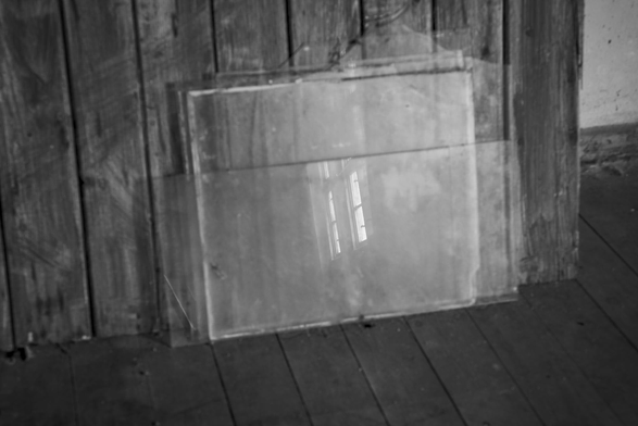 Zwei große, rechteckige Glasscheiben lehnen an einer alten Holzwand auf einem Holzboden, Spiegelung eines Fensters sichtbar.