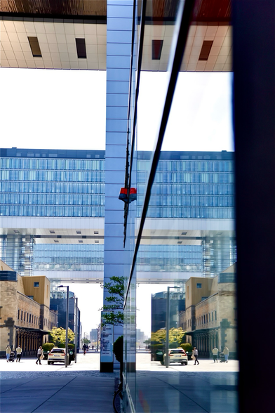 Blick an einer Straße entlang.  Auf der linken Seite befinden sich ein Backsteingebäude und dahinter ragen zwei sogenannte Kranhäuser, benannt nach ihrer Bauform, raus. Die Kranhäuser haben jede Menge Fenster. Auf der Straße laufen zwei Fußgänger mit Kinderwagen, daneben steht ein Auto, in das Personen einsteigen. Diese Szene wird circa in der Mitte des Bildes an einer sehr großen Glasfront genauso gespiegelt. Das es eine Spiegelung ist, erkennt man am ehesten an einer roten Signalleuchte, die eng an der Scheibenfront sitzt und doppelt wirkt, da sie direkt daneben gespiegelt wurde. 