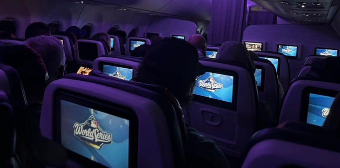 4+ rows of in-flight airplane passengers are watching the same thing on their seatback screens: a blue background with a “World Series” logo. The cabin lights are dimmed purple, so the screens dominate the image. There are two holdouts who seem to have their attention on other types of screen content. [Somewhere over eastern Canada, November 2025]