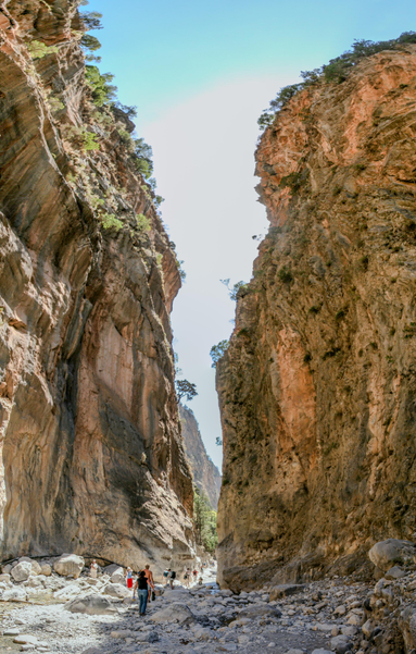 A group of people walking through a narrow, rocky canyon with towering cliff walls on either side under a clear blue sky.