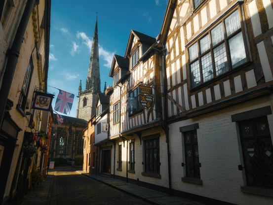 A historic narrow street in Shrewsbury is framed by timber-framed and whitewashed stone buildings on either side, their dark wooden beams contrasting sharply against cream-coloured walls, with small-paned windows and dark doorways visible along the street level. The distinctive tall spire of St. Alkmund's Church rises prominently in the background, its pointed Gothic architecture piercing a brilliant blue sky dotted with wispy white clouds. In the foreground on the left, the Prince Rupert Hotel displays hanging heraldic signs and decorative banners, including the Union Jack flag, whilst traditional pub signs hang from wrought-iron brackets. The warm sunlight casts long shadows across the cobbled street, highlighting the architectural details of these medieval buildings and creating a picturesque, timeless scene typical of England's historic market towns.
