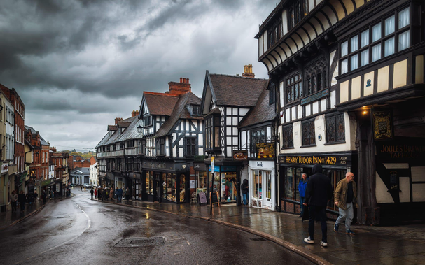 A wet street slopes gently downhill along Wyle Cop in Shrewsbury, lined with rows of striking timber-framed buildings dating back centuries, their distinctive black wooden beams creating bold geometric patterns against cream and white-rendered walls. The Henry Tudor Inn, dated 1429, stands prominently amongst the shopfronts on the right-hand side, with various independent retailers and galleries occupying the ground floors of these historic buildings. A handful of pedestrians move along the damp pavement beneath an overcast, dramatic sky filled with grey and white clouds, whilst the wet surface reflects the warm glow of shop windows and street lighting. The street recedes into the distance with rows of further historic buildings flanking both sides, creating a tunnel-like perspective that emphasises the medieval character and charm of this remarkably well-preserved Shropshire town centre, with glimpses of the surrounding hillsides visible beyond the buildings.
